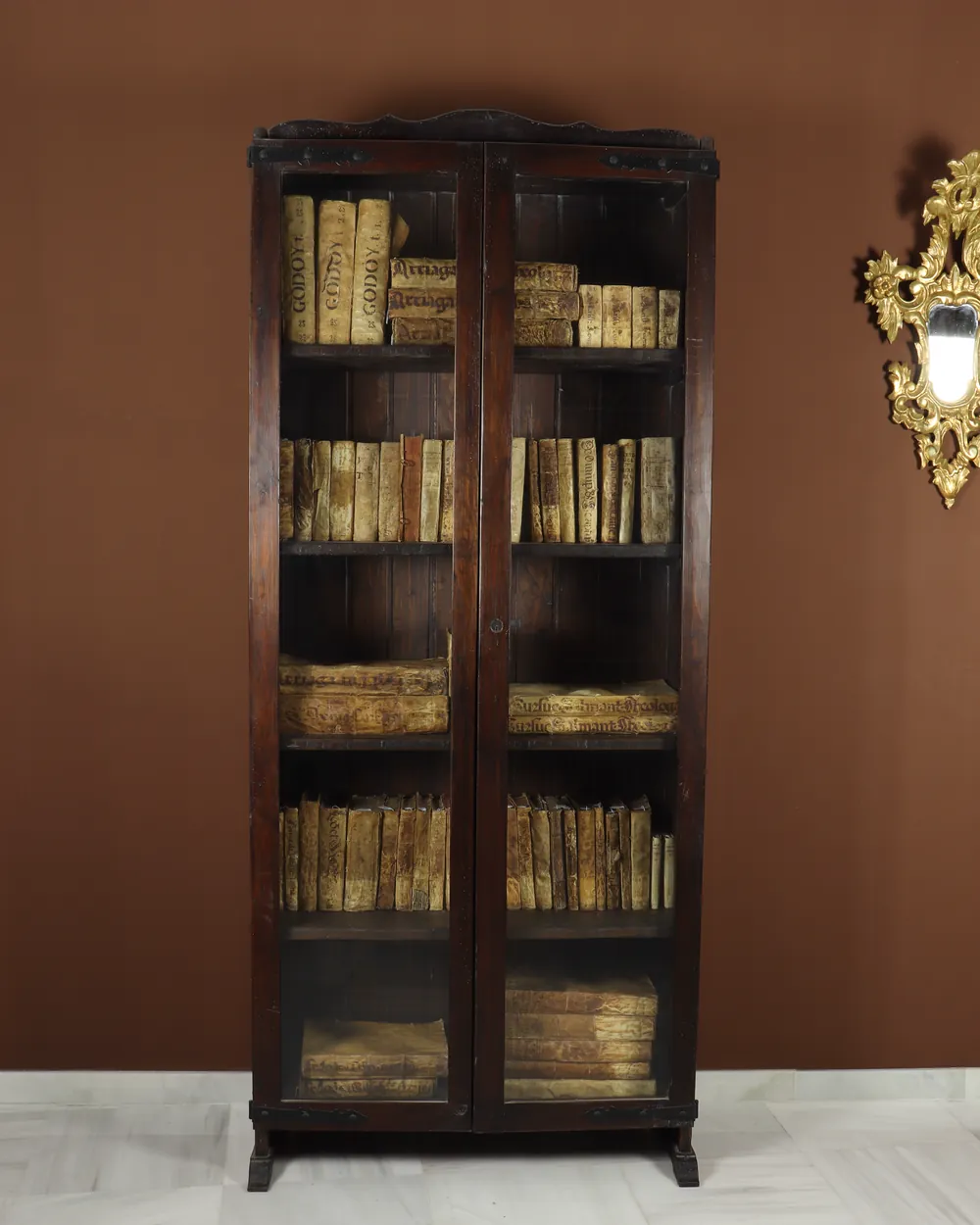 Un armario de madera oscura con las puertas de cristal cerradas, lleno de libros antiguos encuadernados en pergamino, ante una pared marrón y junto a un espejo de marco dorado