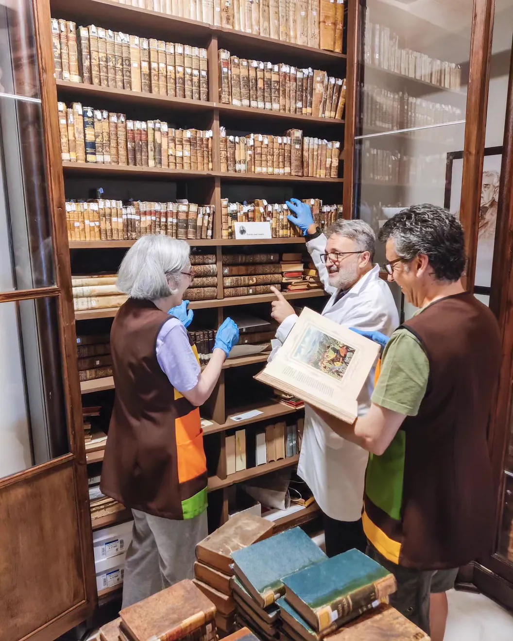 Tres personas con guantes azules ante un armario de libros encuadernados en pergamino; una de ellas, con bata blanca, sostiene abierto un libro grande con un grabado en color