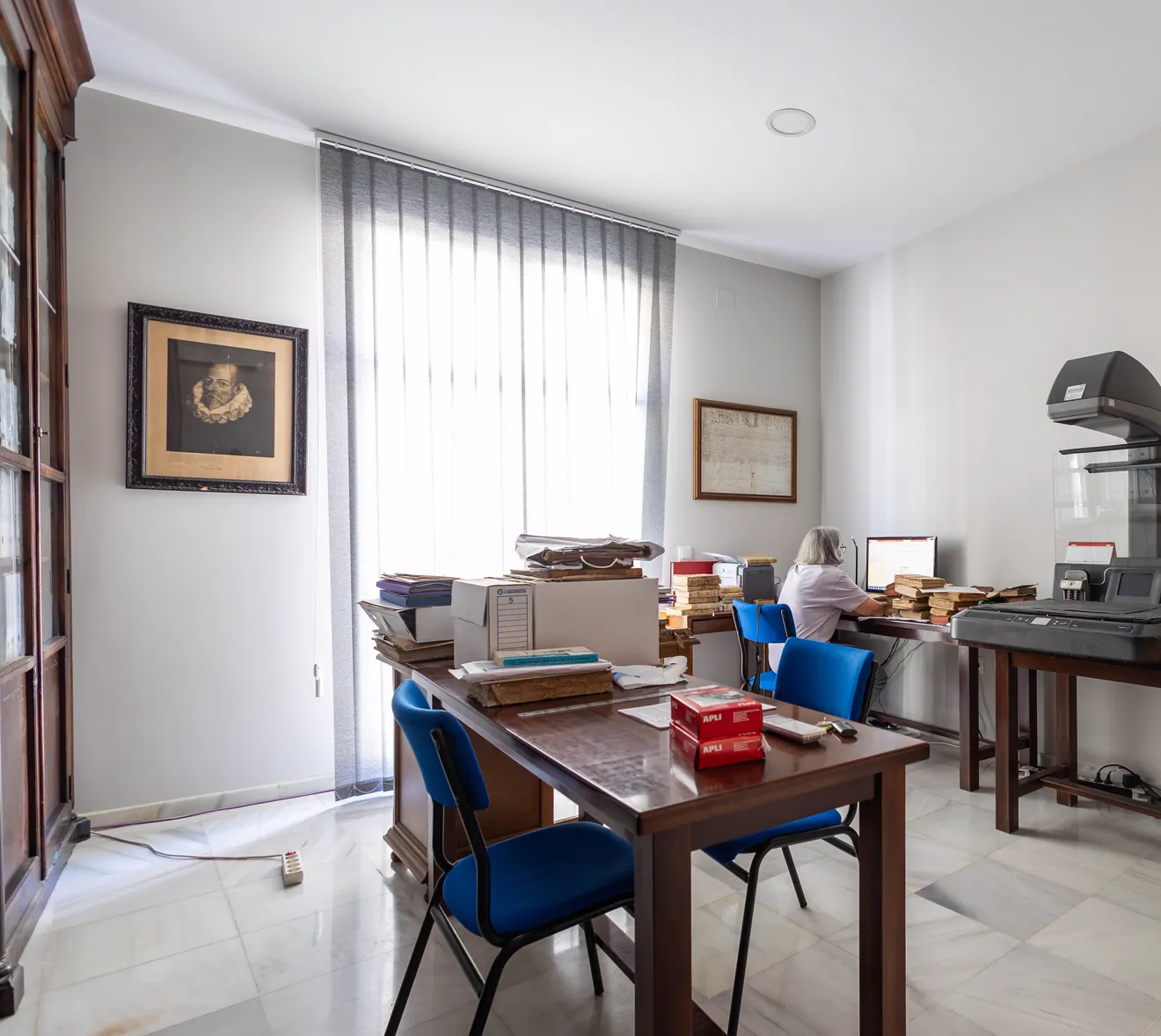 Un cuarto de trabajo con una mesa llena de cajas y libros, una mujer al ordenador al fondo, un retrato antiguo enmarcado en la pared y, a la derecha, un escáner de libros