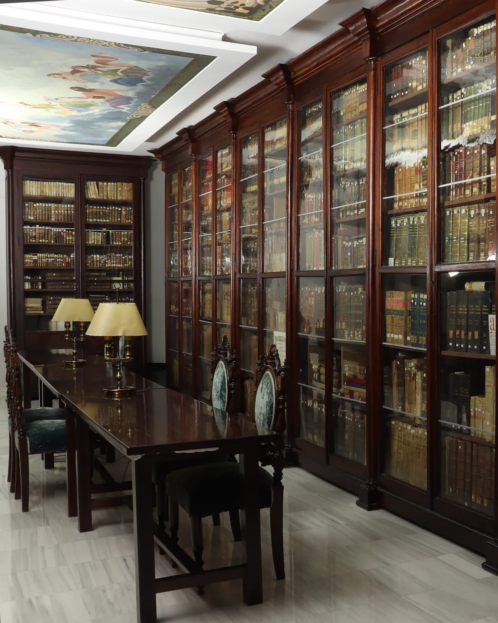 Una sala con armarios de madera oscura y puertas de cristal llenos de libros antiguos y una mesa de lectura con lámparas de pantalla dorada, bajo un techo con pinturas de nubes y figuras