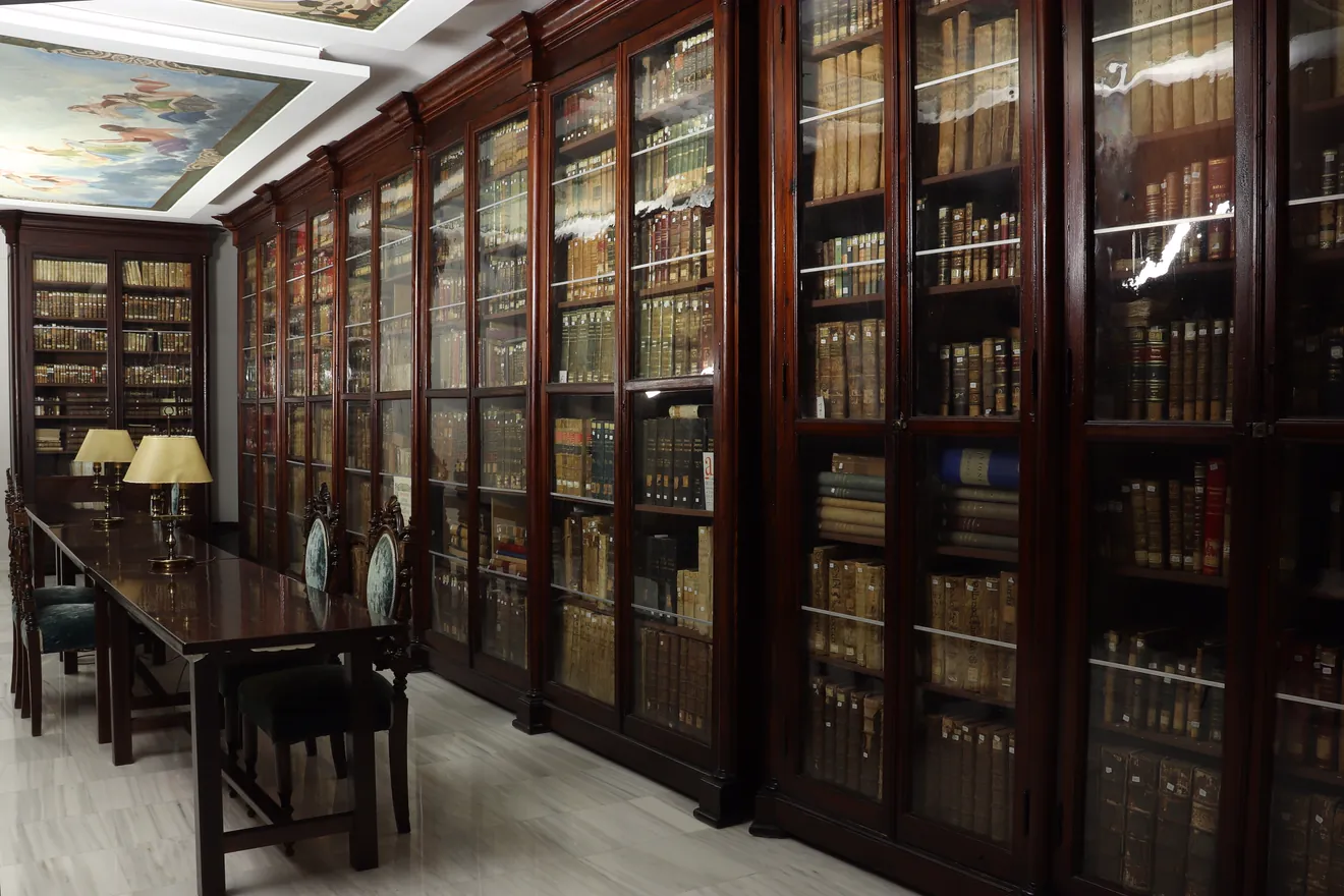 Una sala larga con armarios de madera oscura y puertas de cristal llenos de libros antiguos, una mesa de lectura con lámparas de pantalla dorada y sillas tapizadas, bajo un techo con pinturas de nubes y figuras