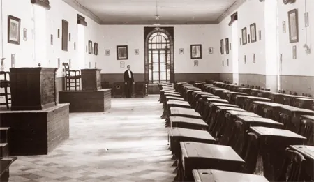 Fotografía antigua de un aula larga con pupitres de madera en filas y retratos en las paredes