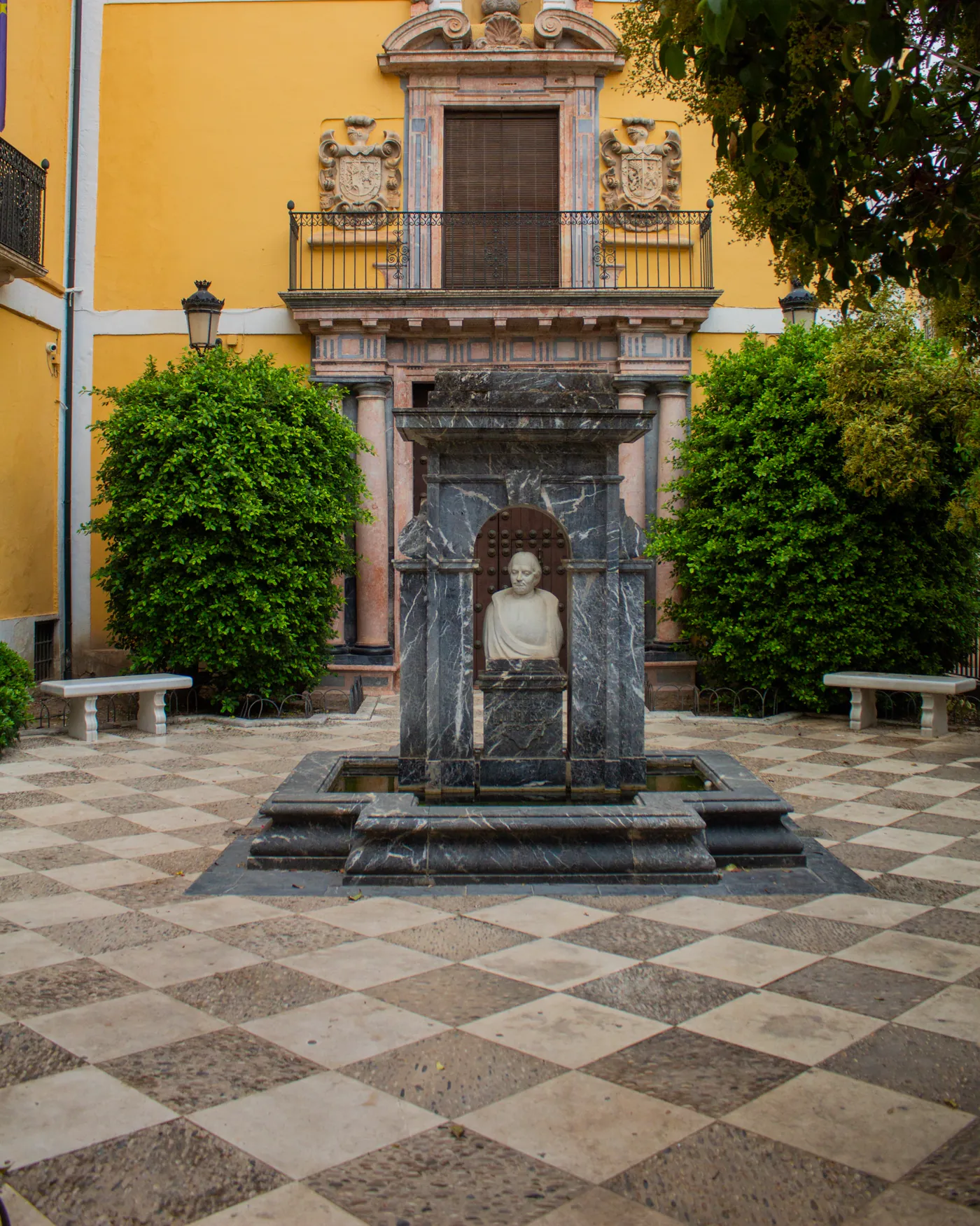 La fuente del fundador, con su busto en piedra, delante de la portada barroca del edificio, entre setos y bancos de la plaza de Aguilar y Eslava