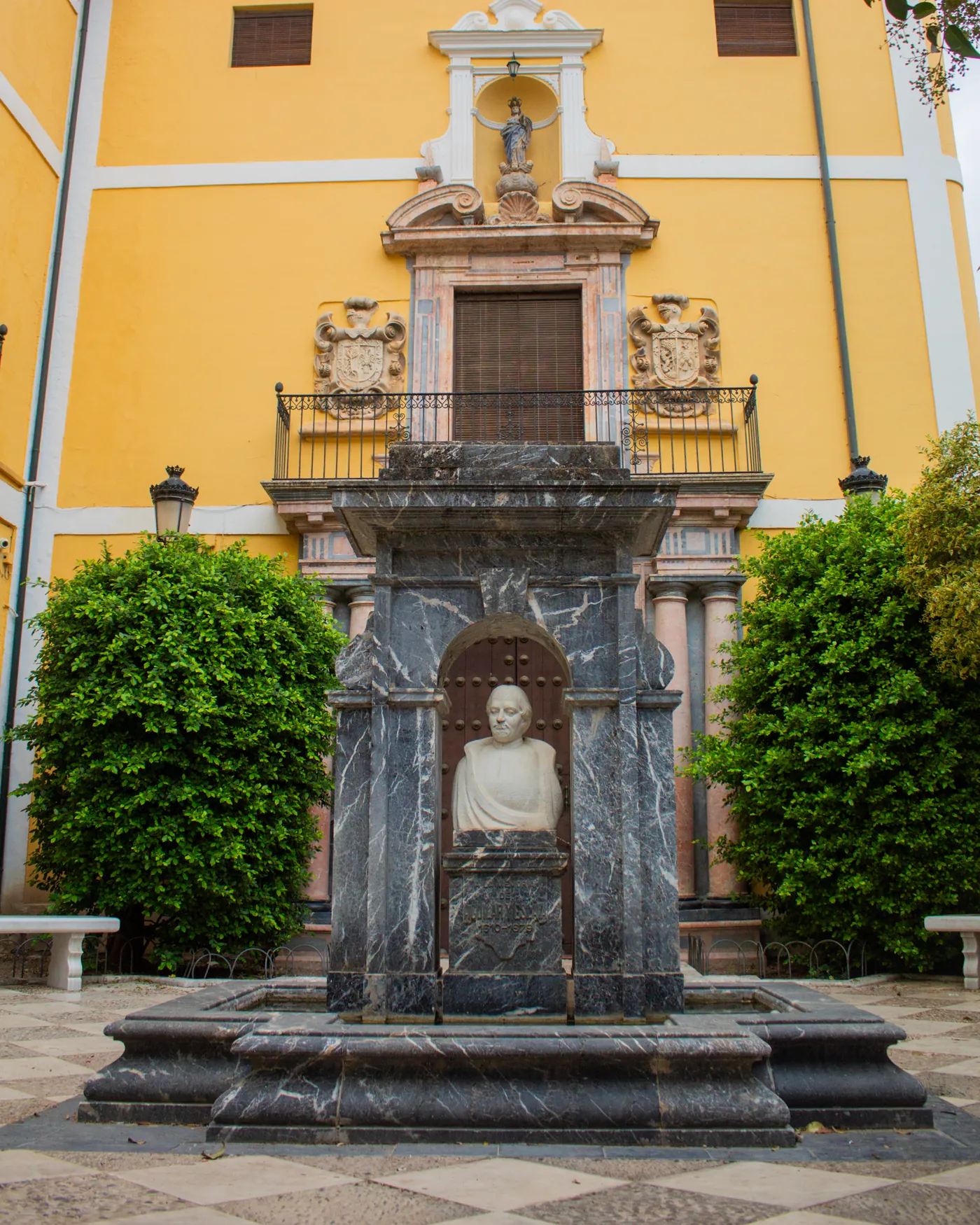 La portada barroca del edificio, en mármol rojo y negro, con su balcón entre dos escudos y la hornacina de la Purísima; delante, la fuente en honor del fundador con su busto en piedra, en la plaza de Aguilar y Eslava